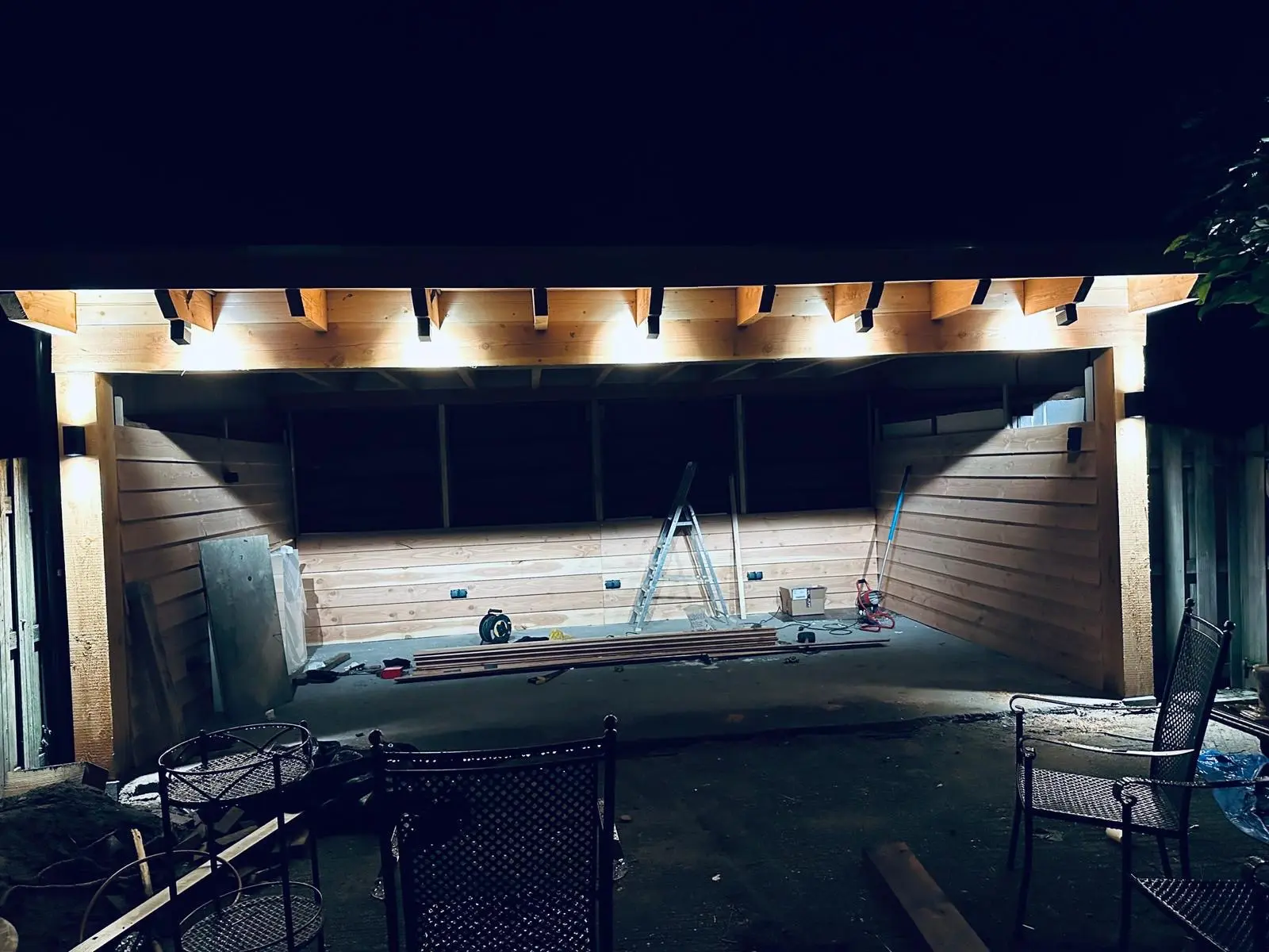 Houten wandbetimmering en dakconstructie van een overkapping, verlicht met inbouwspots in de avond